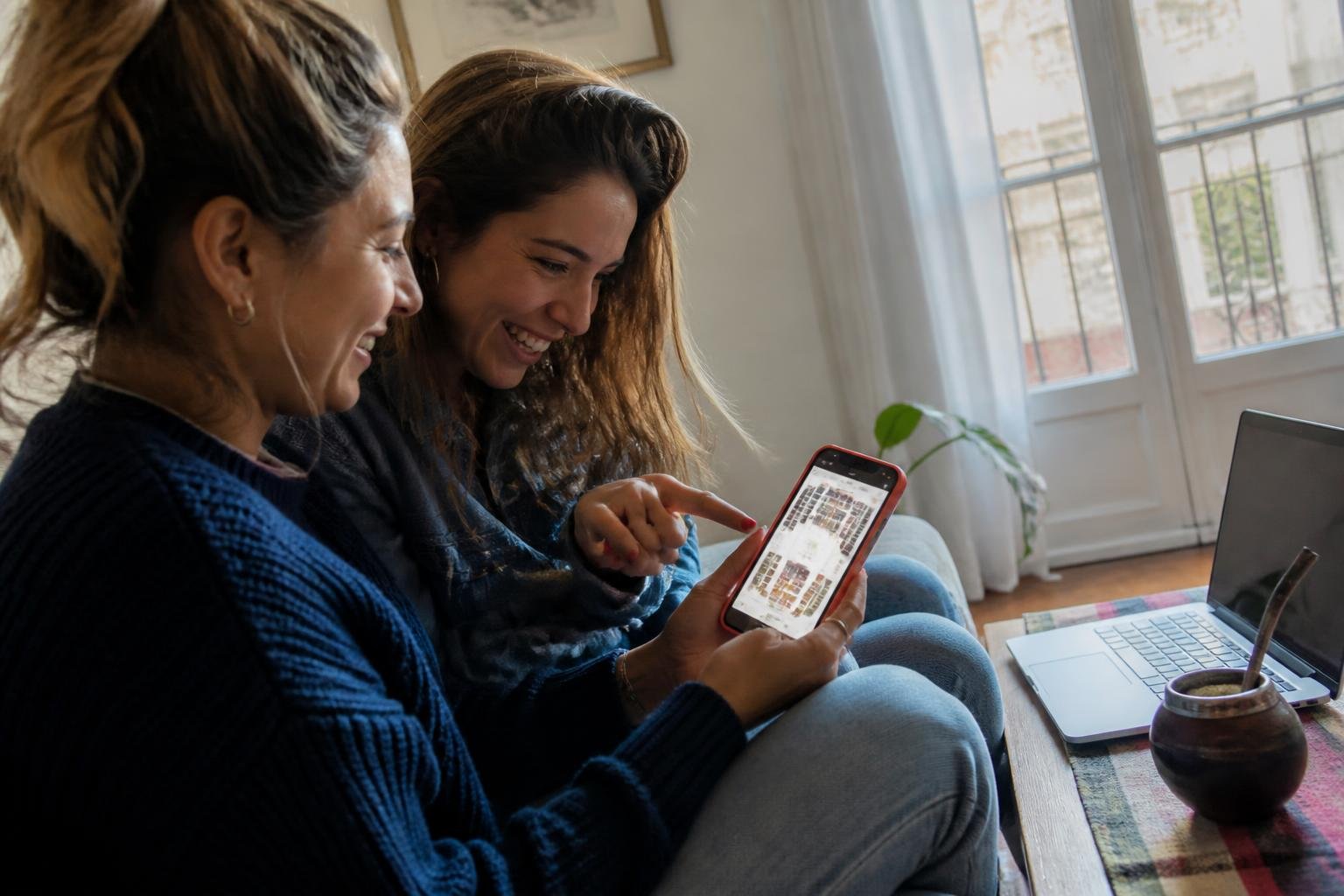 Dos amigas comprando entradas de Broadway desde el celular en el living de su casa, con un mate en la mesa
